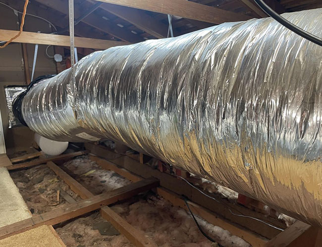 A large, metallic duct stretches through an attic, resting on wooden beams, with insulation visible on the floor beneath.