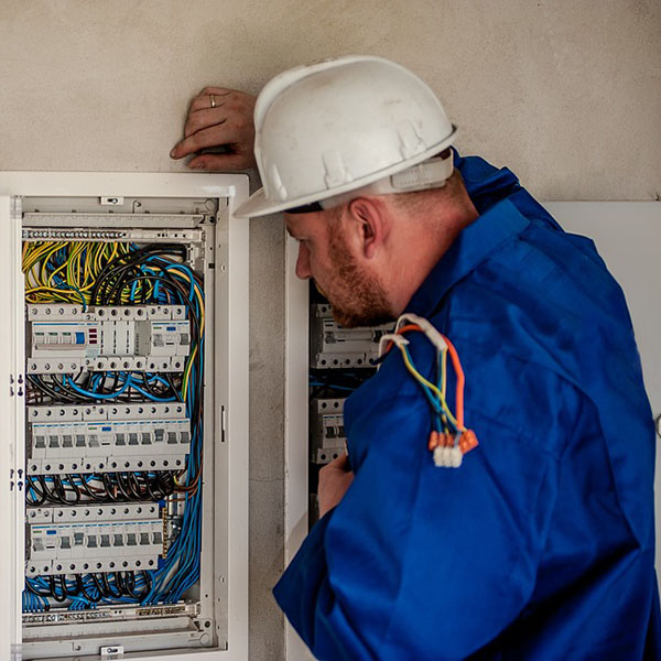 An electrician in a blue jacket examines a wall-mounted electrical panel filled with colored wires and circuit breakers.