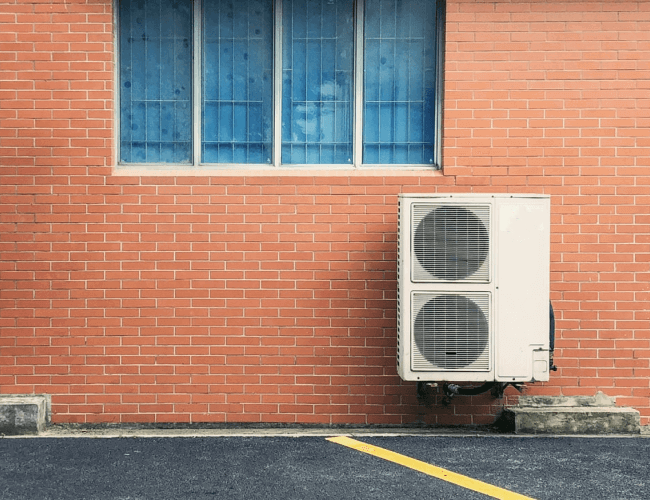 Air conditioner mounted on the side of a brick building, blending with the structure's exterior.