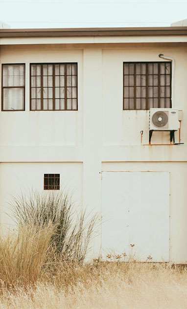 A white building facade with two windows and an air conditioning unit, surrounded by tall grass and a closed door below.