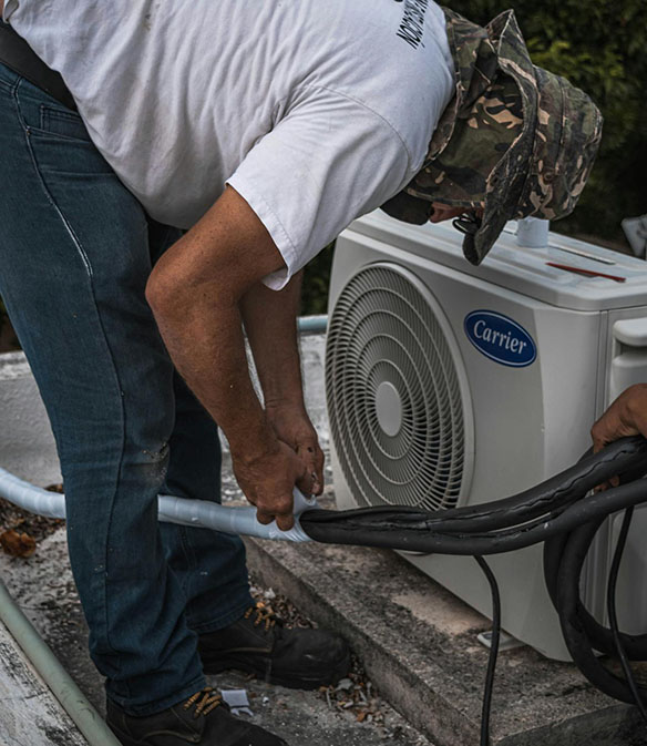 A technician in a camo hat connects tubing to a Carrier air conditioning unit on a rooftop, focusing intently on the task.